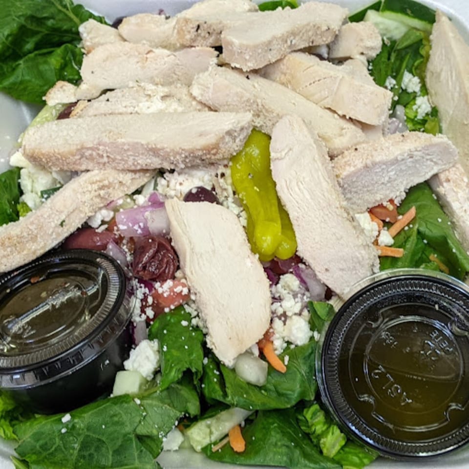 A large salad with sliced chicken, feta cheese, olives, and a side of dressing.