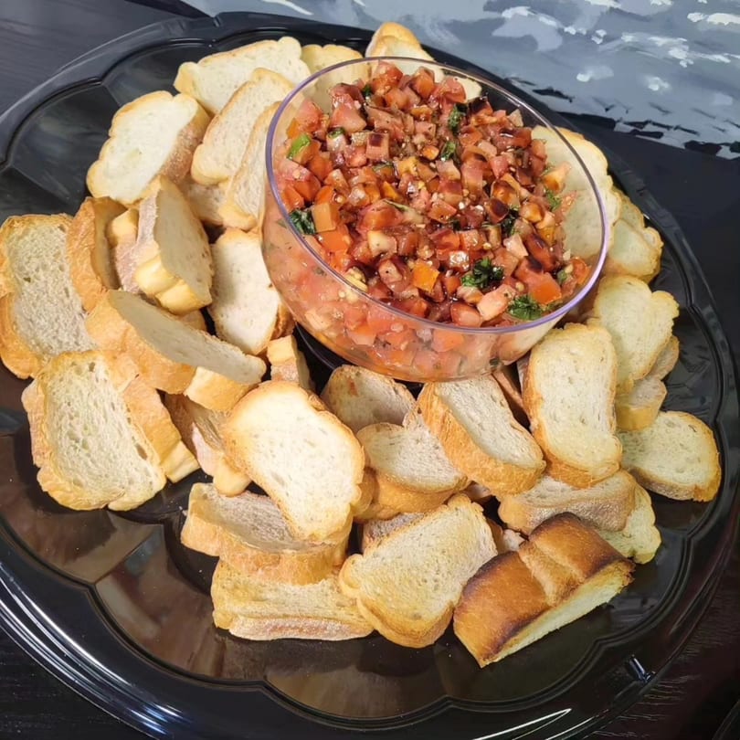 A black platter filled with toasted baguette slices surrounding a glass bowl of fresh bruschetta.