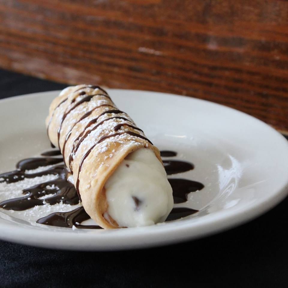 A Pana Roma Pizza & Pasta cannoli, drizzled with chocolate sauce and powdered sugar on a white plate.