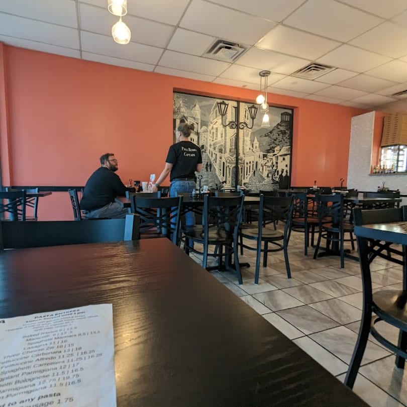 A Pana Roma Pizza & Pasta dining room with orange walls, black tables, and a server assisting a customer.